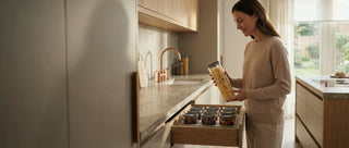 Lifestyle-Fotografie einer Frau in einer minimalistischen Luxusküche, die ein 1,7L HATTY HAYS Borosilikatglas über einer perfekt organisierten Schublade mit dem modularen Aufbewahrungssystem präsentiert.